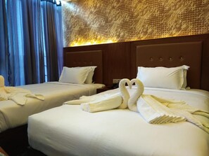A hotel room with two beds, each with white linens and pillows. There are two swan-shaped towels on the beds. The room has a wooden headboard and a textured wall above the beds.