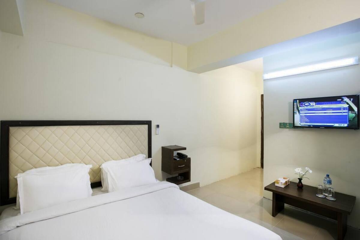 Ein Hotelzimmer mit einem Bett, einem an der Wand montierten Fernseher, einem kleinen Tisch mit einer Vase und einer Flasche sowie einem Nachttisch.