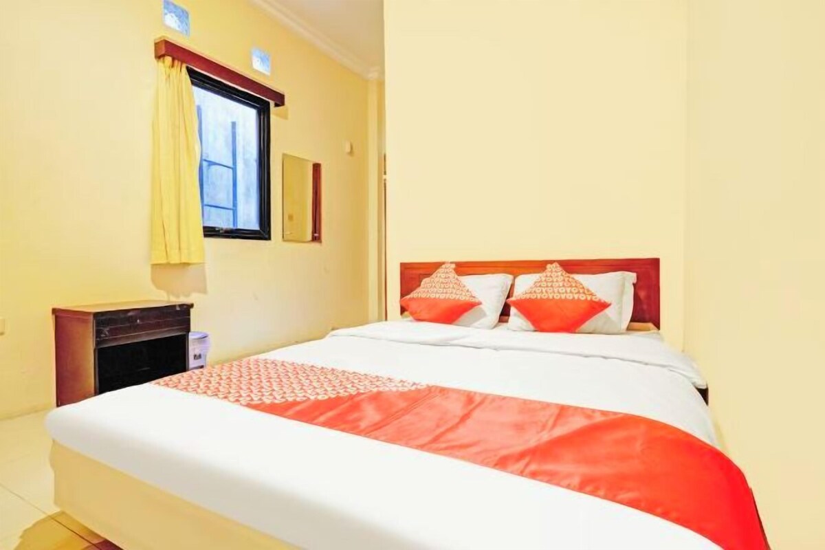 Ein Hotelzimmer mit einem Bett, einem Nachttisch, einem Fenster mit gelben Vorhängen und einem Spiegel an der Wand.
