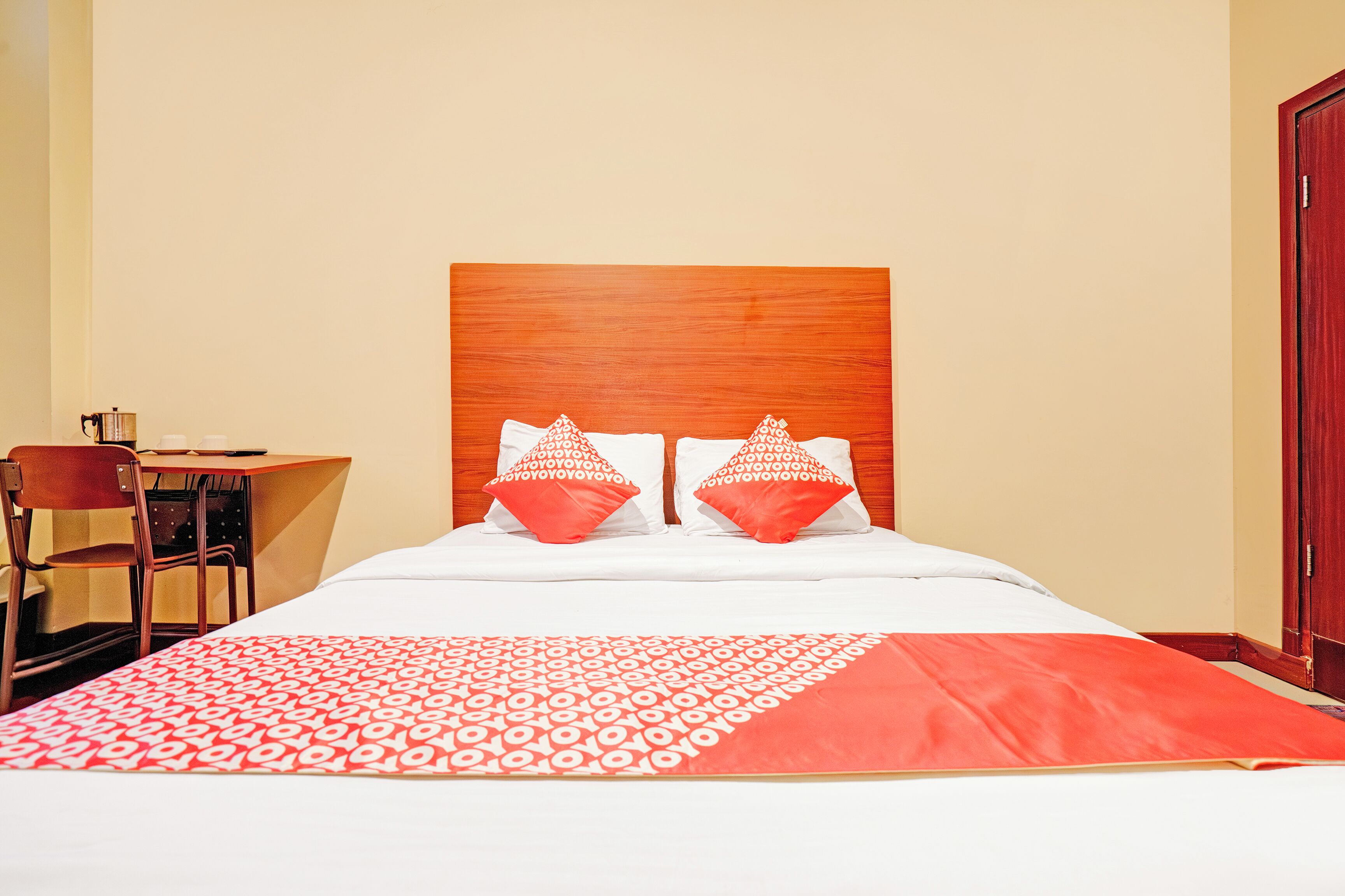 A hotel room with a wooden headboard, a bed with white and red bedding, a desk with a chair, and a wooden door.