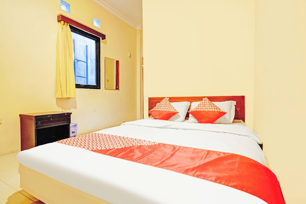 A neatly made bed with a red and white bedspread, a wooden nightstand, a window with yellow curtains, and a mirror on the wall.
