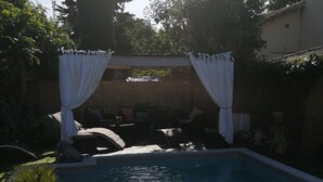 Une piscine dont la surface de l’eau est illuminée par le soleil, entourée d’un espace de détente abrité avec des chaises et une table, et un pot de fleurs en premier plan.