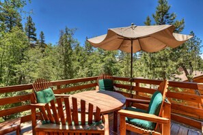A wooden deck with a round table and chairs, an umbrella, and a forested backdrop.