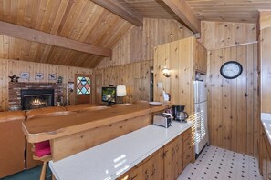 Un interior de madera con chimenea, televisor y una zona de cocina con refrigerador y reloj.