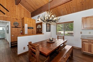 A wooden interior with a dining table, benches, a chandelier, and a view through a window. - Eagle Point Cabin Backing to 7 Acres of Preserve! HOT TUB!  A/C! PET FRIENDLY! Walk to Lake & Shuttles! (Big Bear Lake)