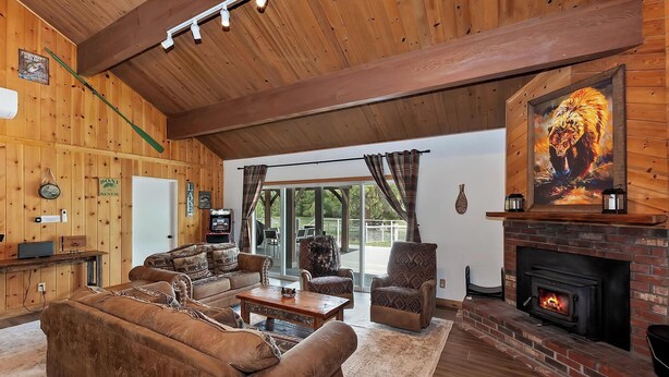 A cozy cabin interior with a fireplace, wooden walls, and a large window overlooking a deck. - Eagle Point Cabin Backing to 7 Acres of Preserve! HOT TUB!  A/C! PET FRIENDLY! Walk to Lake & Shuttles! (Big Bear Lake)