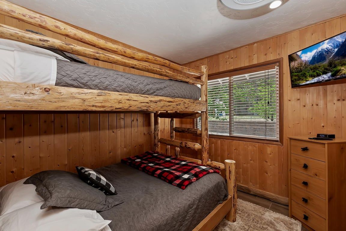 A bunk bed with a wooden headboard, a bedside table, a drawer unit, and a window with blinds.