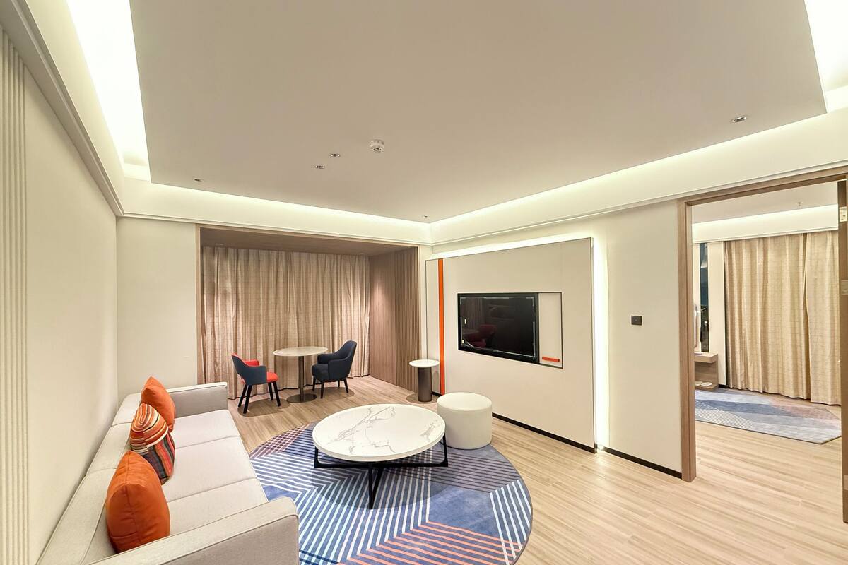 A modern hotel room with a sofa, two chairs, a coffee table, a TV, and a large window.