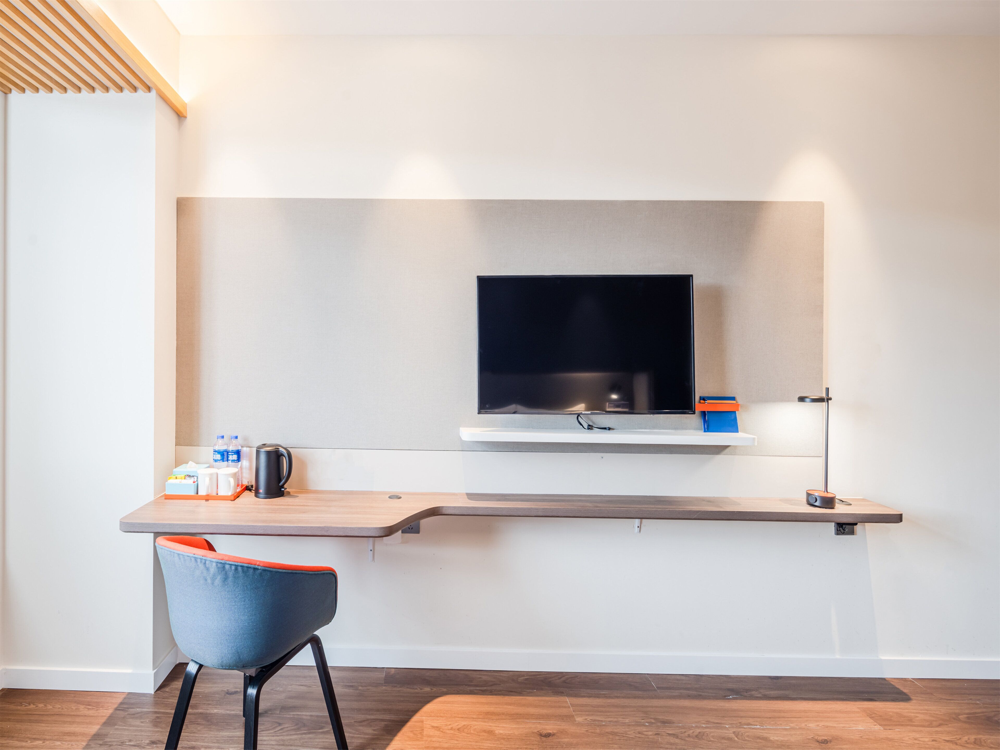 Une chambre moderne avec un téléviseur à écran plat fixé à un mur blanc, un bureau en bois avec une chaise et une petite étagère contenant divers objets.