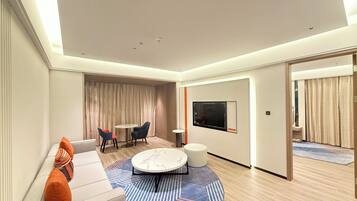 A modern hotel room with a sofa, two chairs, a coffee table, a TV, and a large window.