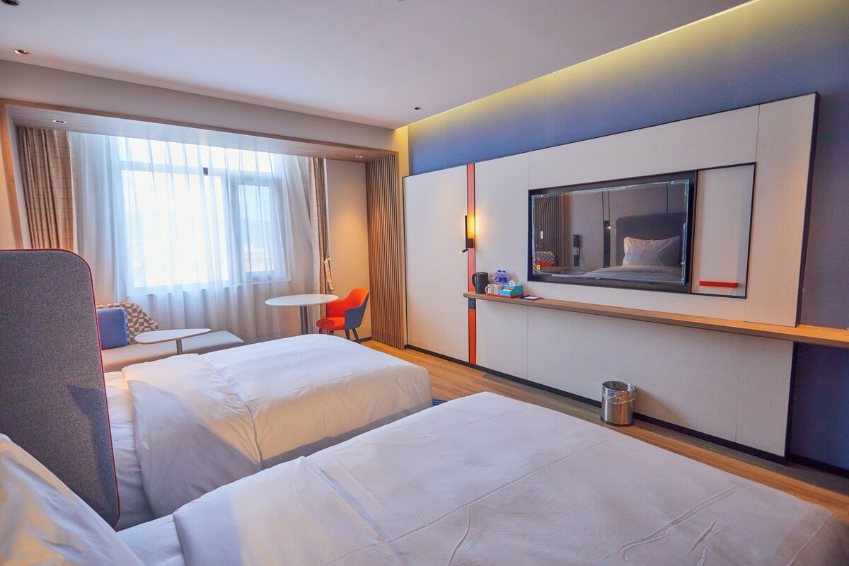 A hotel room with two beds, a small table, a chair, a TV, and a window with curtains.