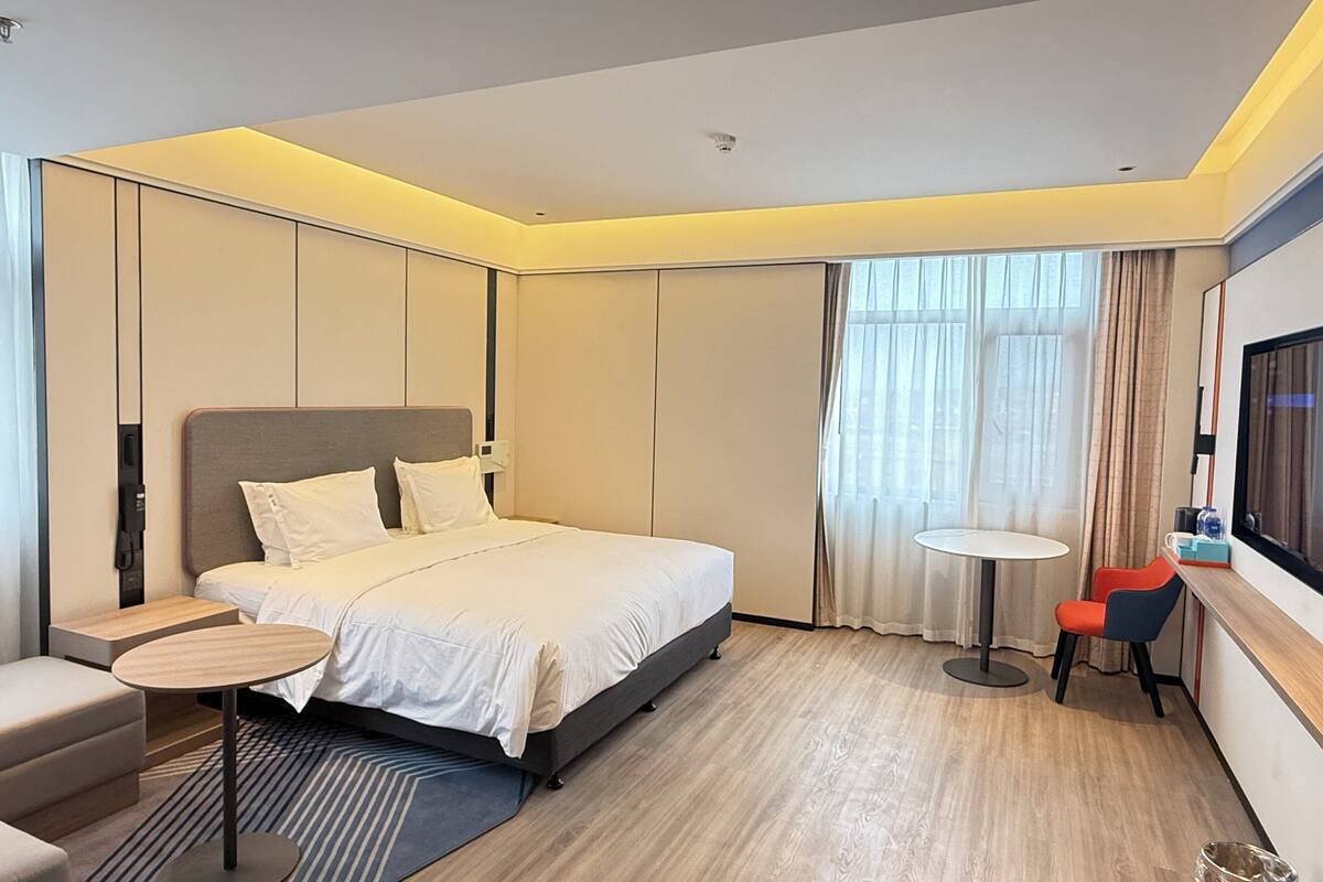 A modern hotel room with a large bed, a small round table, a chair, a TV, and a window with curtains.