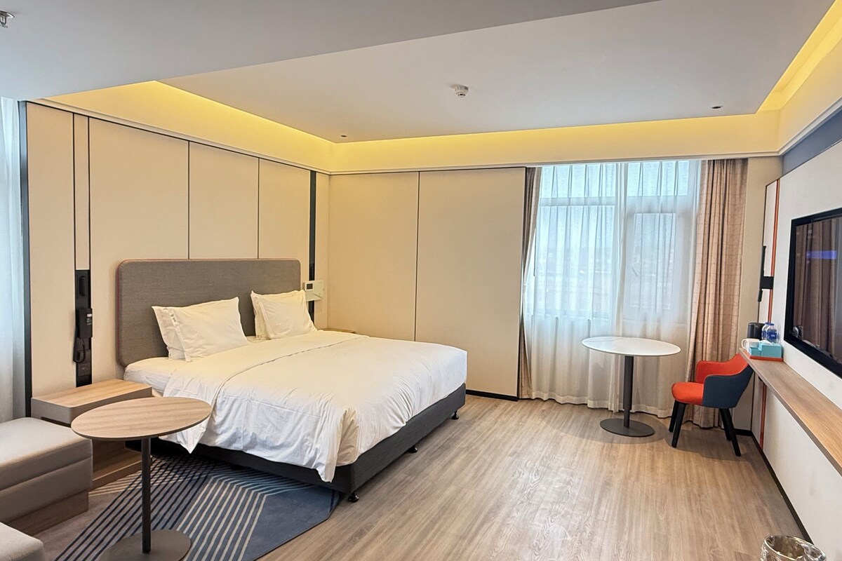 A modern hotel room with a large bed, a small round table, a chair, a TV, and a window with curtains.