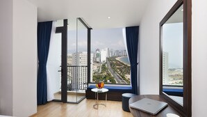 A modern hotel room with a large window offering a city view, a small table with a laptop, a cup, and a bowl of fruit. - Crown Hotel Nha Trang (Nha Trang)
