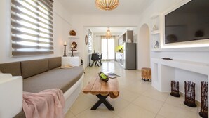 A modern living room with a sofa, a wooden coffee table with a laptop and a bowl of fruit, a hanging light fixture, and a TV mounted on the wall. - Rocks Naxian Villa, private Pool, by NaxosVibe (AGIA ANNA, NAXOS)