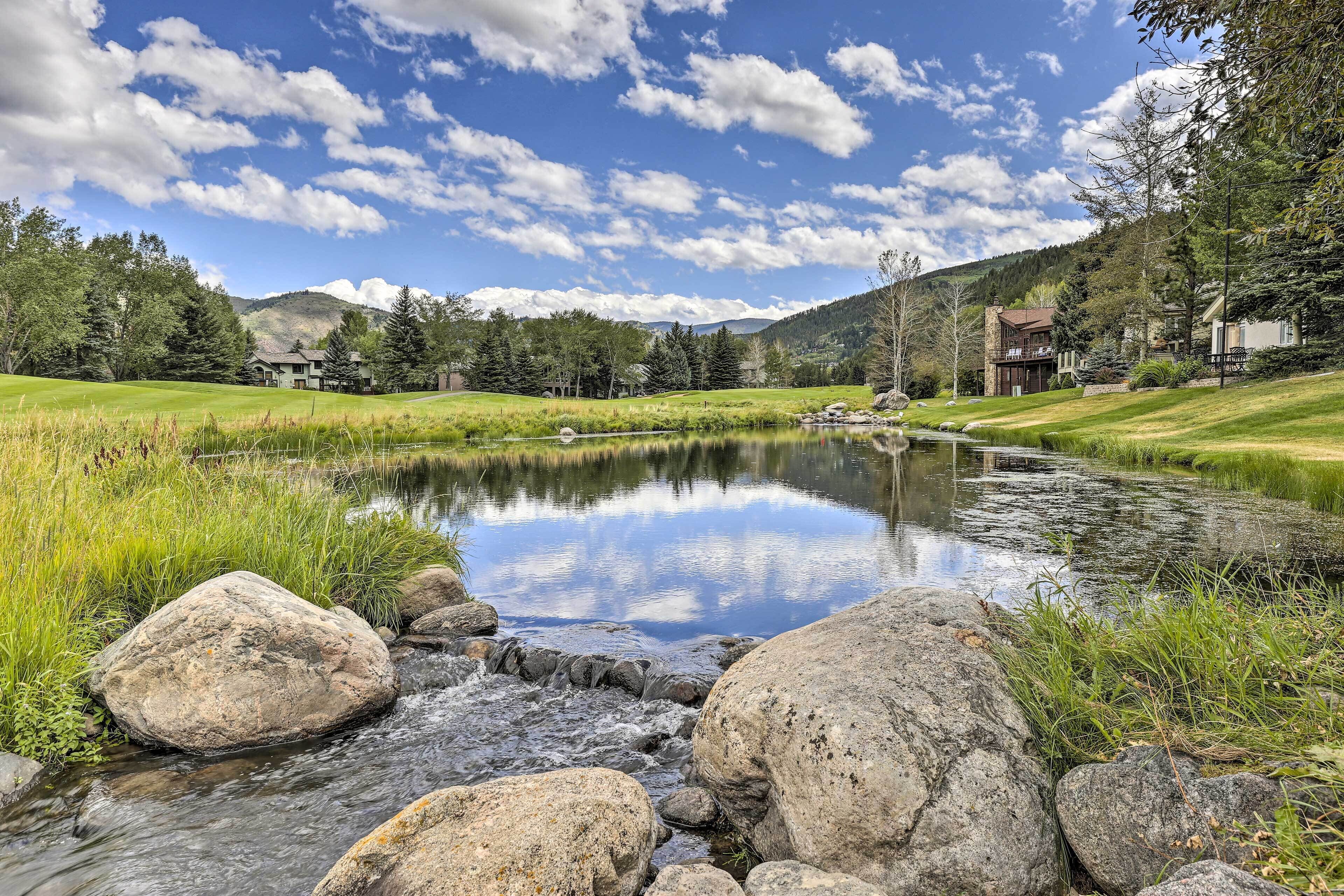 A serene lakeside view with a small stream, surrounded by lush greenery and mountains in the background.