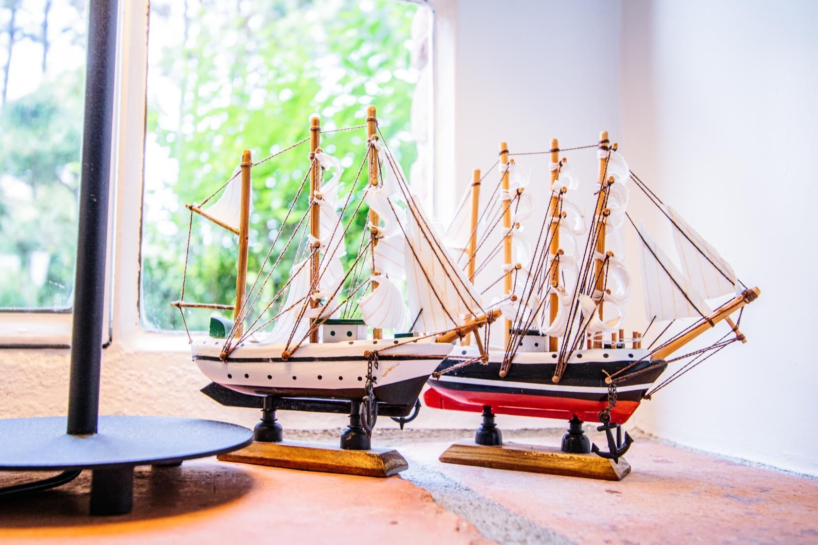 Two model sailing ships on display stands near a window with a view of greenery.
