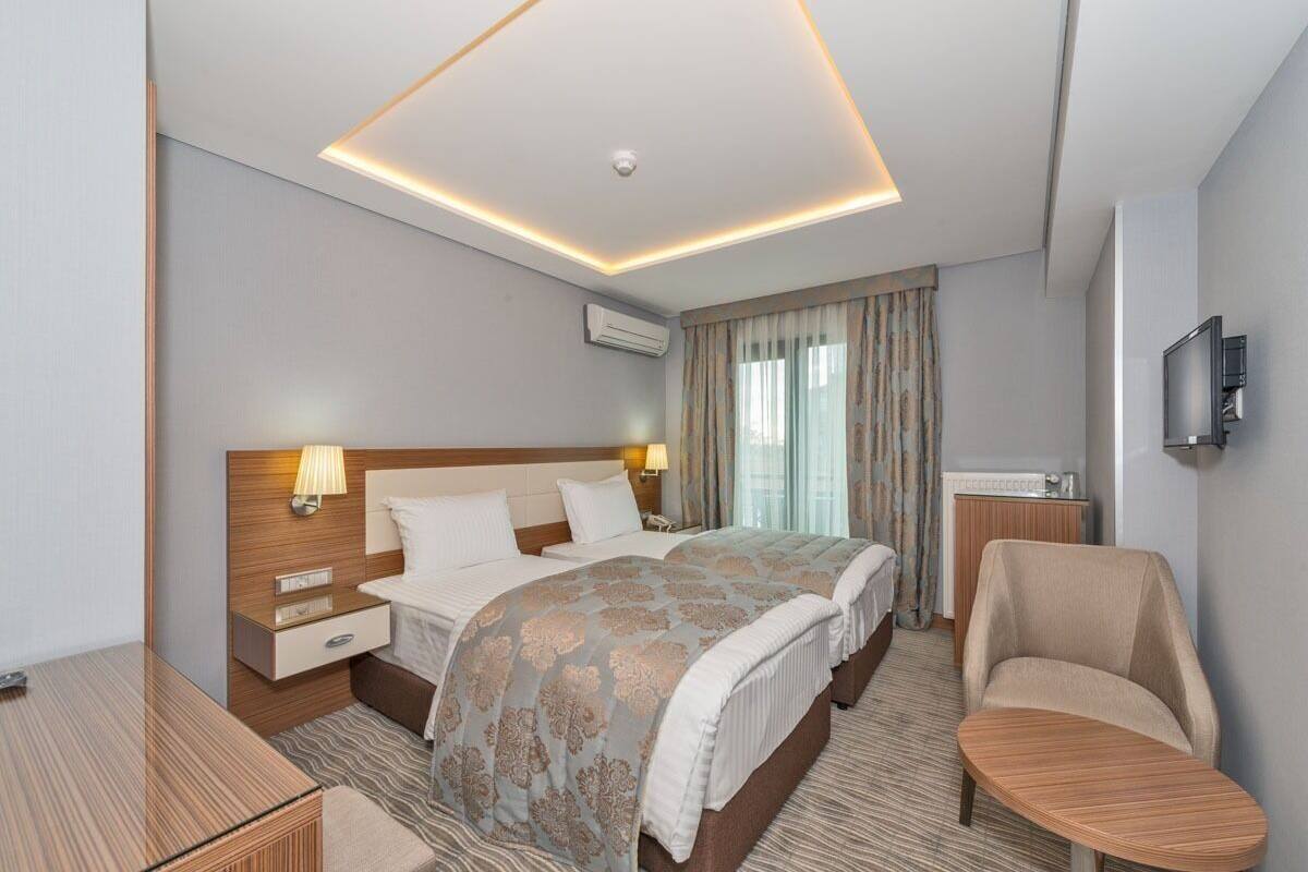 A hotel room with a bed, two bedside tables, a desk, a chair, an air conditioning unit, a television, and a window with curtains.