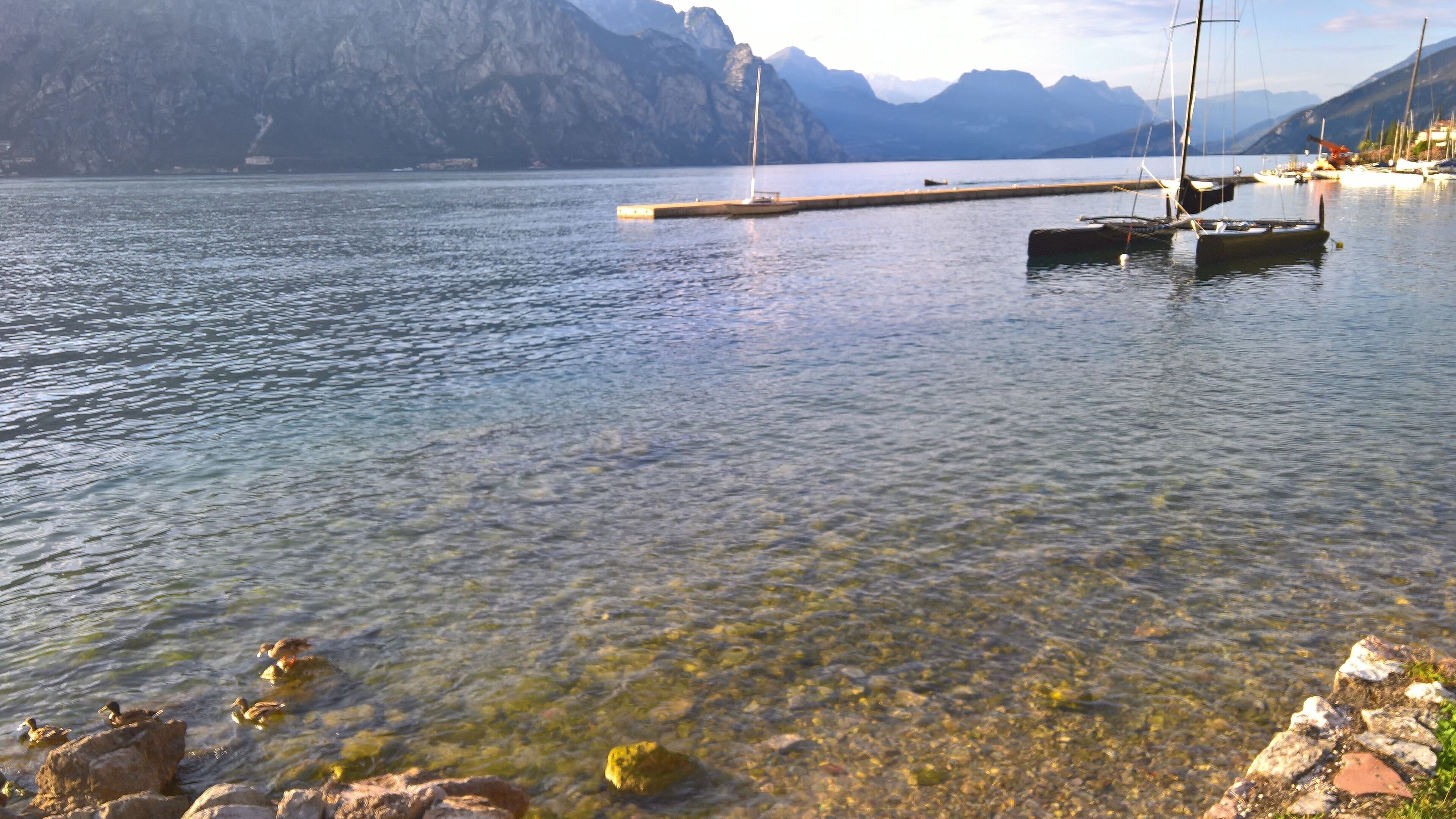 Um lago calmo com águas cristalinas, um cais e barcos. Montanhas visíveis ao fundo.