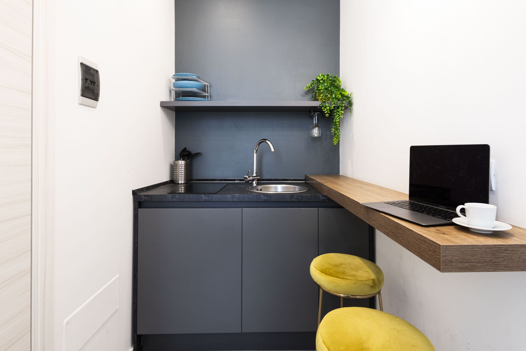 A compact kitchen with a sink, a countertop, and a laptop on a wooden shelf.