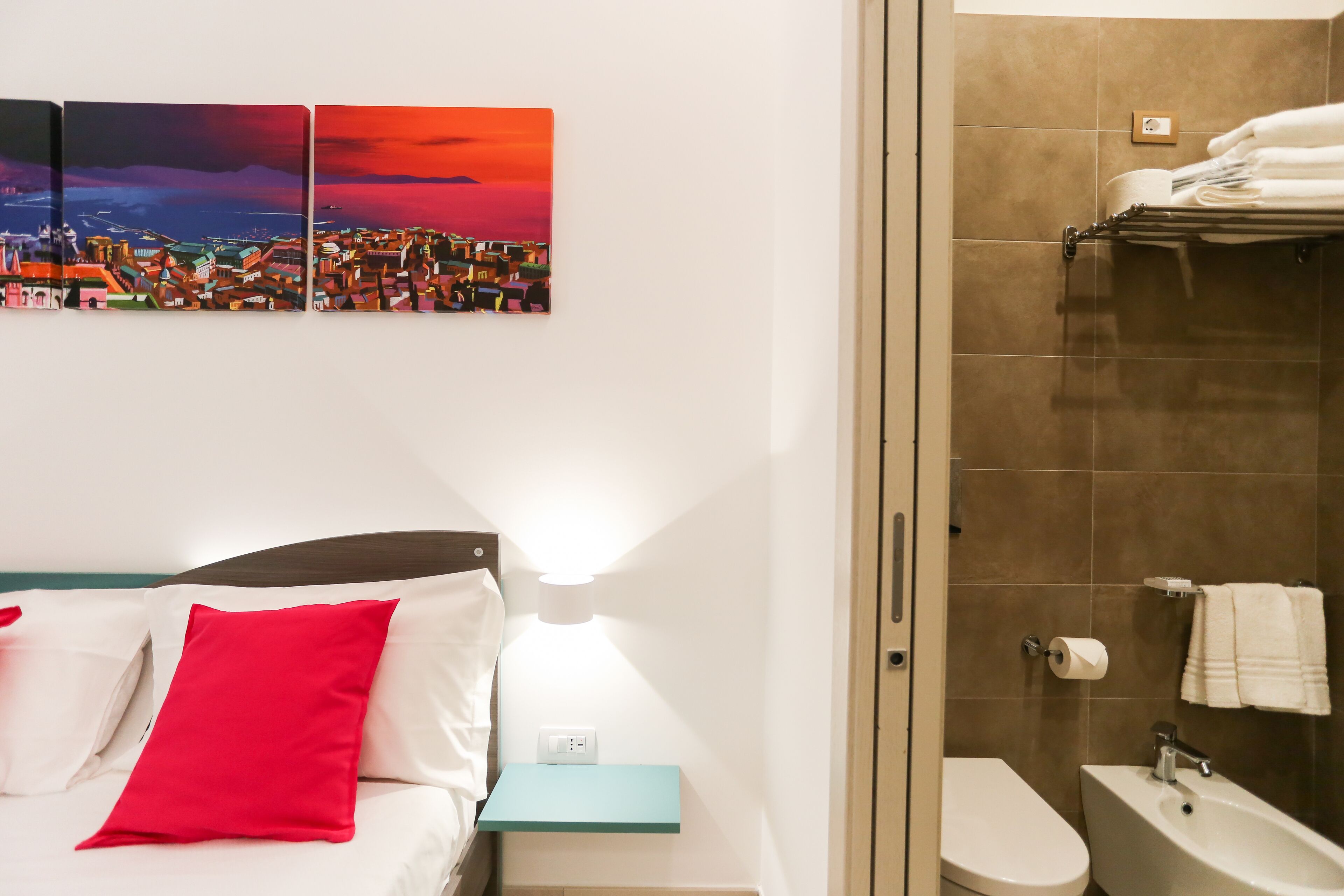 A hotel room with a bed, a nightstand, a wall-mounted lamp, a bathroom with a toilet, sink, and towel rack.