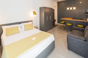 A modern hotel room with a large bed, a grey sofa, a small kitchen area with stools, and a wardrobe.