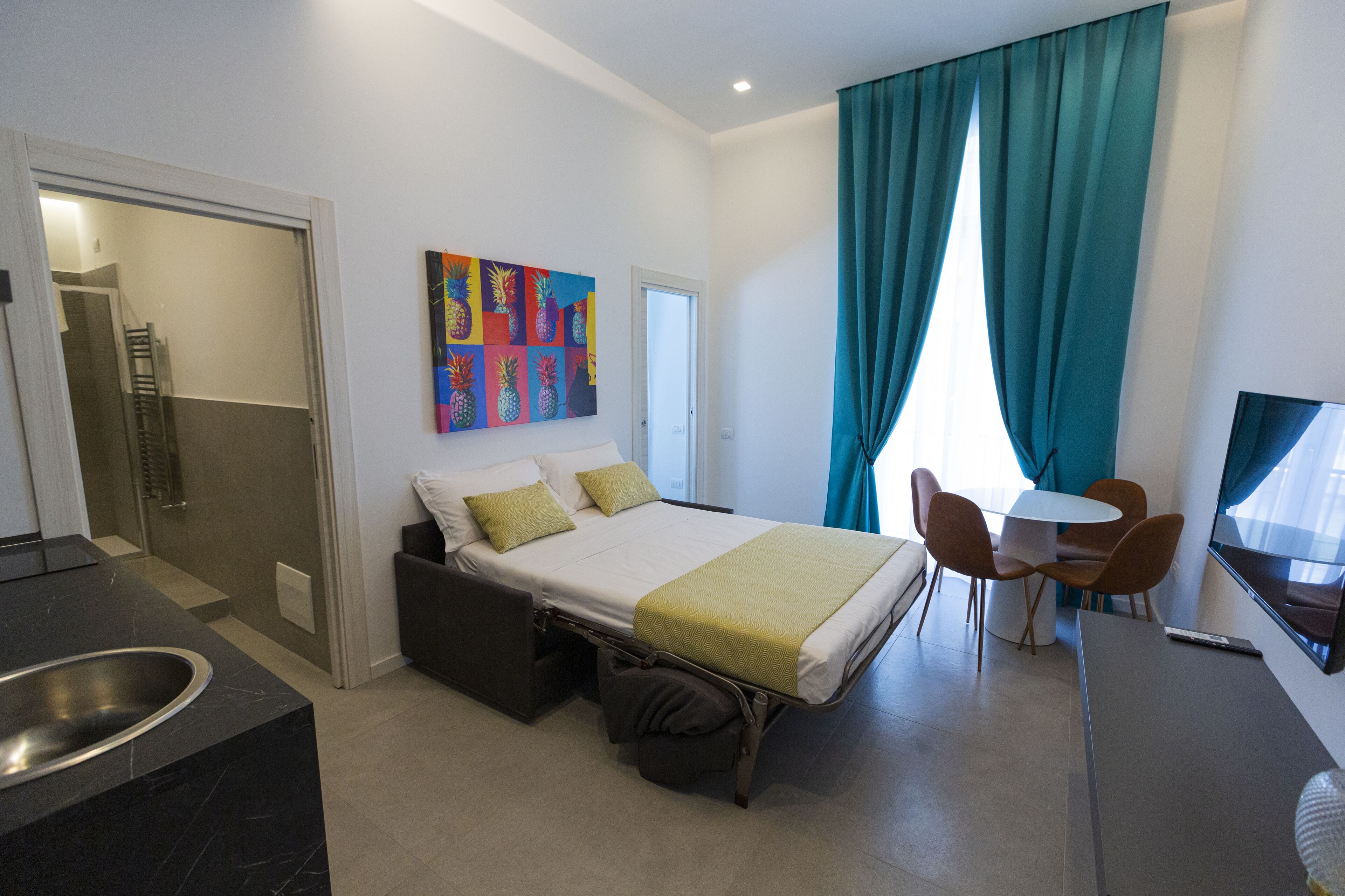 Quarto de hotel moderno com cama, uma pequena escrivaninha, televisão e banheiro com chuveiro e vaso sanitário.