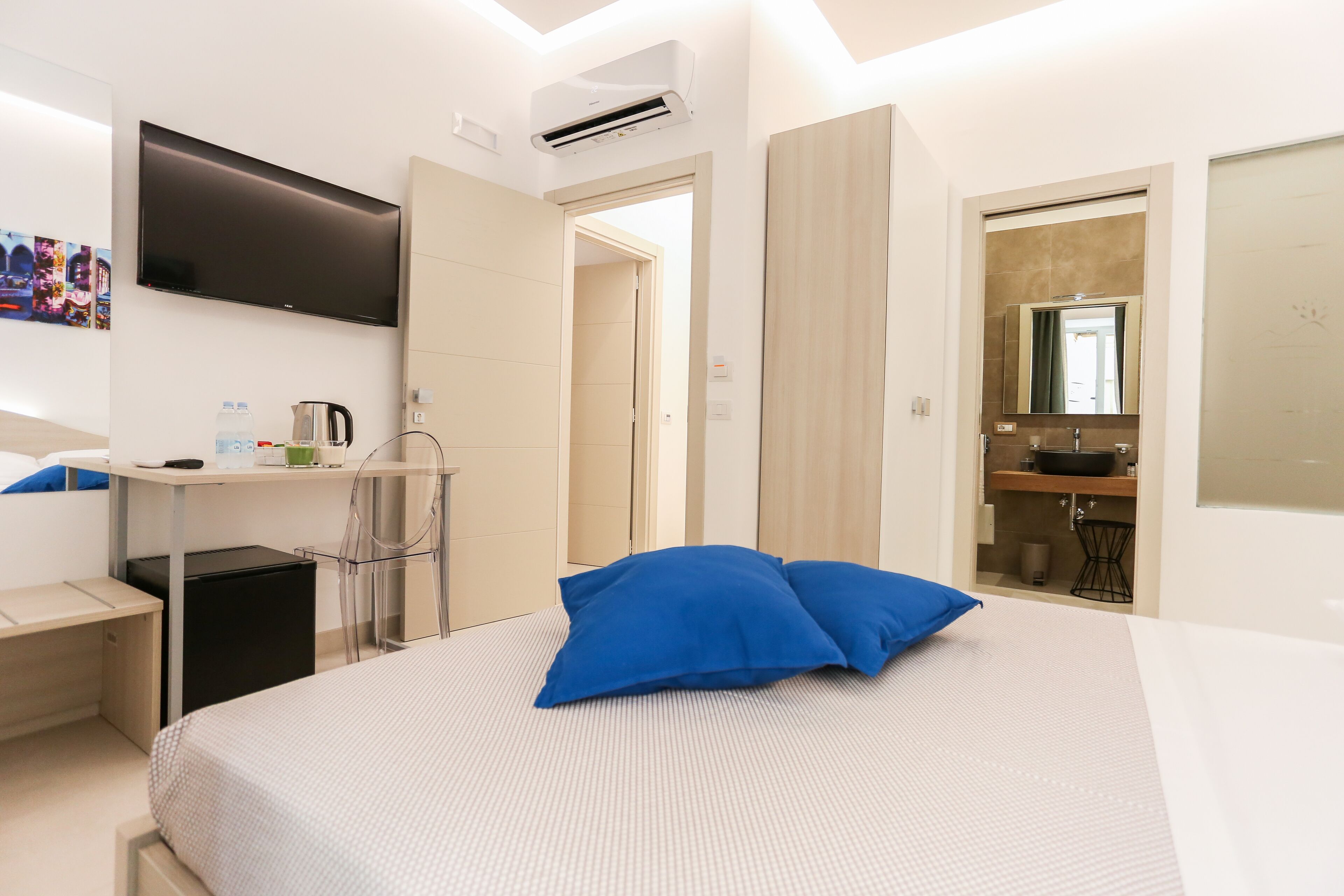 A modern hotel room with a bed, a desk, a flat-screen TV, a mini-fridge, and a bathroom visible through an open door.