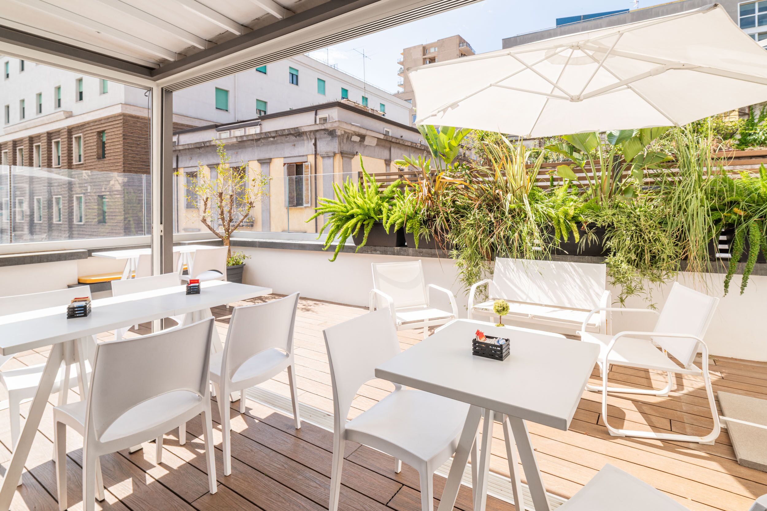 Una terraza al aire libre con mesas y sillas blancas, un paraguas grande y plantas en macetas.