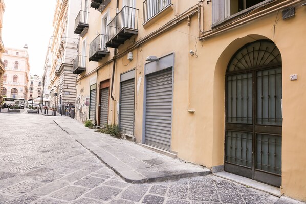 A narrow street with closed shop shutters, a stone pavement, and a yellow building with a large arched window and a barred door.