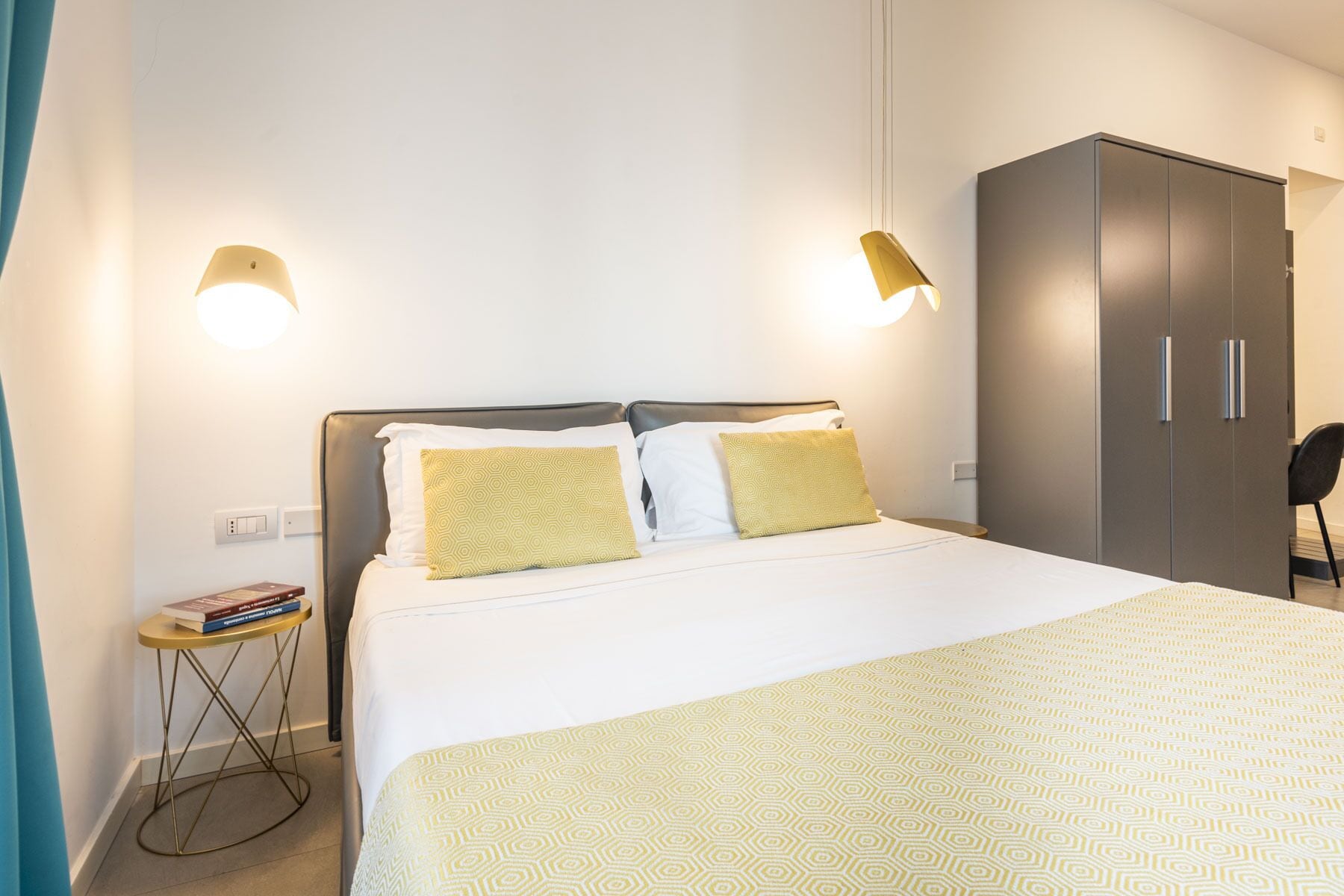 A hotel room with a bed, bedside lamps, a wardrobe, and a small table with books.