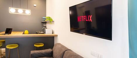A modern living space with a flat-screen TV displaying 'NETFLIX', a kitchenette with a laptop, bar stools, and a sofa.
