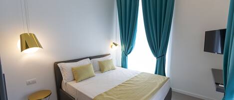 A modern hotel room with a neatly made bed, a small round table, a wall-mounted TV, and teal curtains.