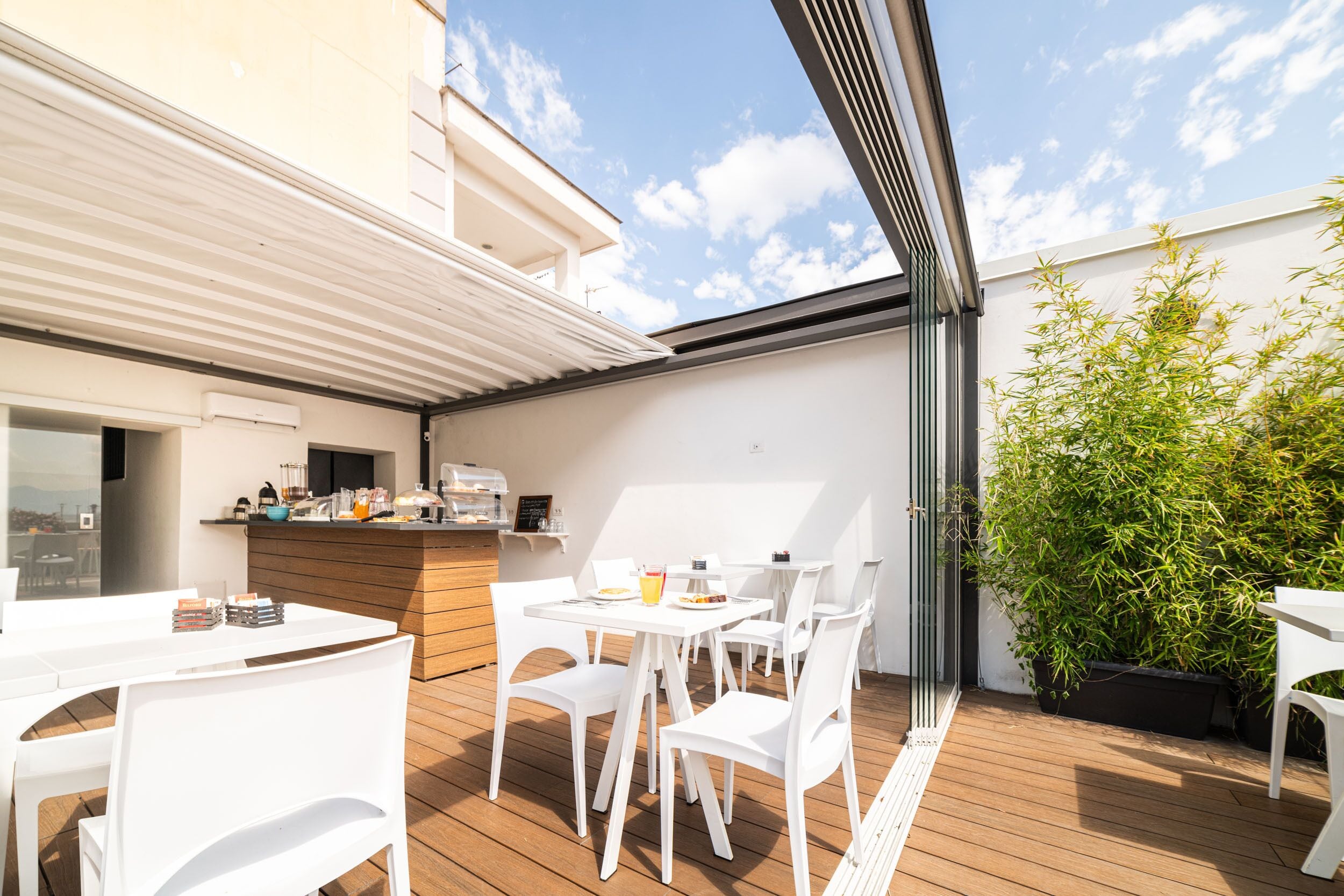 An outdoor patio with white tables and chairs, a wooden deck, a bar area with a counter, and a large potted plant.