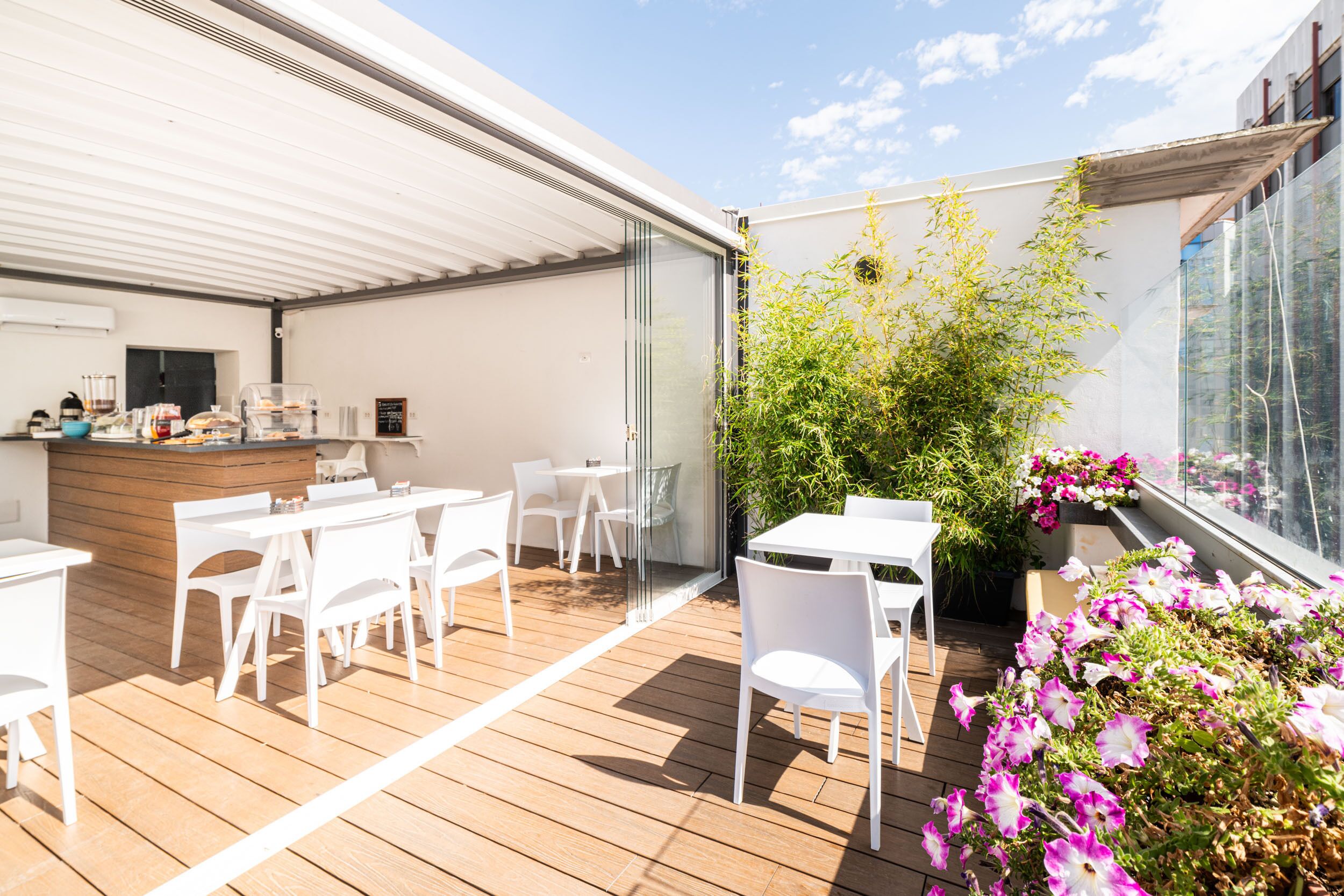 An outdoor patio with white tables and chairs, a wooden counter with a coffee machine, and potted plants with flowers.