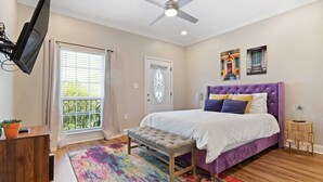 A bedroom with a bed, a television, a ceiling fan, a door, and a window with curtains. - Modern 4BR Mid-City home with heated pool & balcony. Free parking, walk to streetcar & parades. (New Orleans)