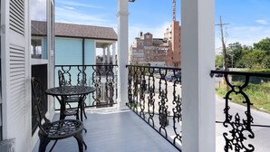 A balcony with a table and chairs, white columns, and a view of buildings in the background. - Modern 4BR Mid-City home with heated pool & balcony. Free parking, walk to streetcar & parades. (New Orleans)