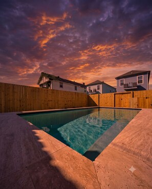 A swimming pool with a wooden deck, surrounded by a wooden fence, and houses in the background under a sunset sky. - Modern 4BR Mid-City home with heated pool & balcony. Free parking, walk to streetcar & parades. (New Orleans)
