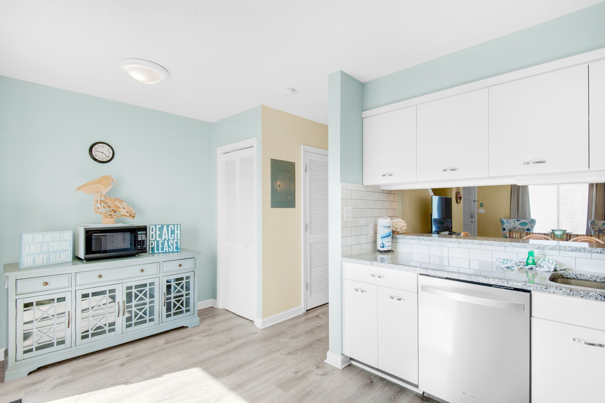 A kitchen with white cabinets, a microwave, a clock, and a sign that reads "The sun and the sand and a drink in my hand."