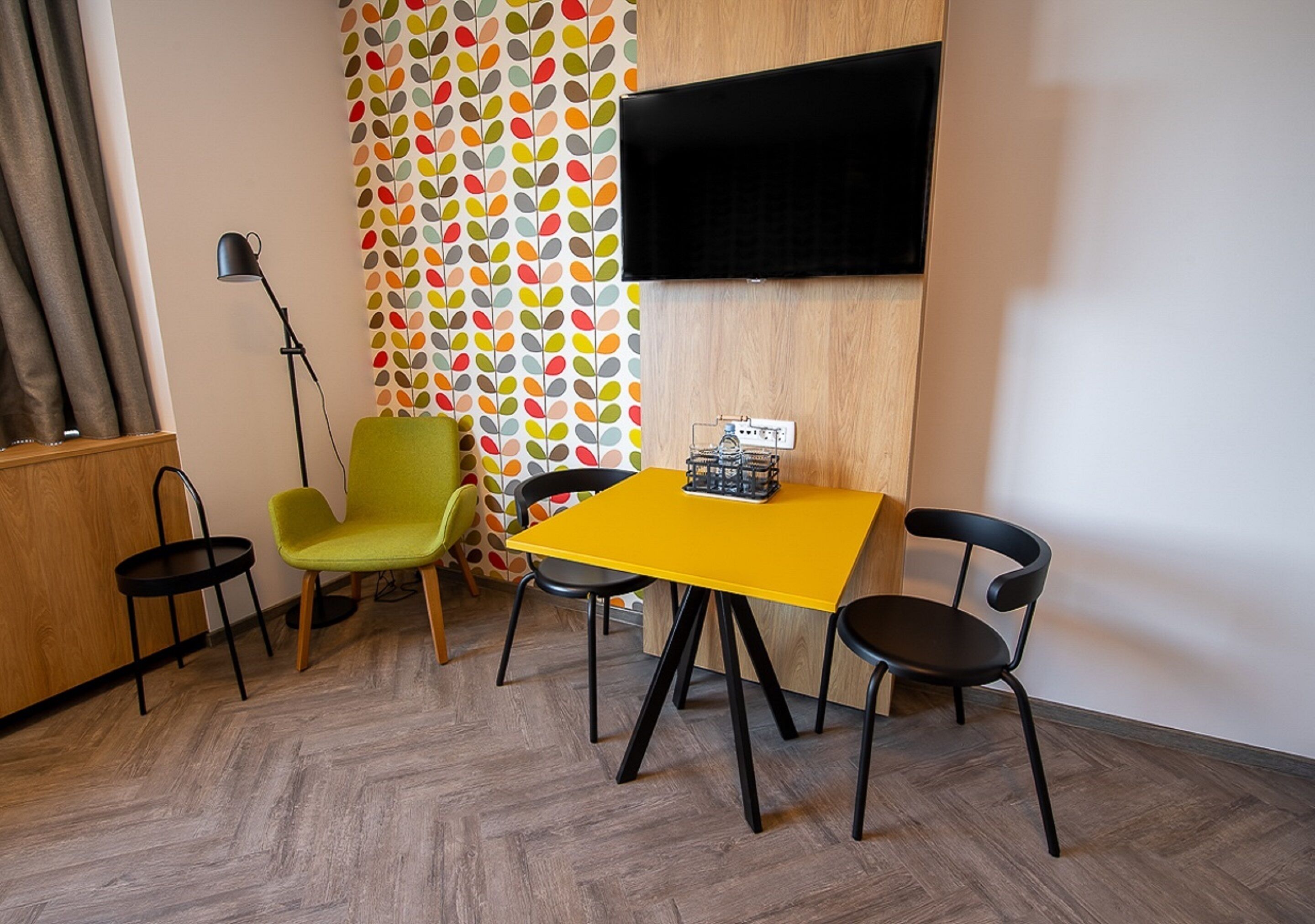 Una habitación con una mesa amarilla, dos sillas negras, un sillón verde, una lámpara de pie negra y un televisor montado en la pared.