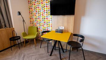 Una habitación con una mesa amarilla, dos sillas negras, un sillón verde, una lámpara de pie negra y un televisor montado en la pared.