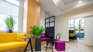 Een moderne woonkamer met een gele bank, een zwarte stoel, een klein tafeltje en een paars voetenbankje. Er staan potplanten en is een houten accentmuur.