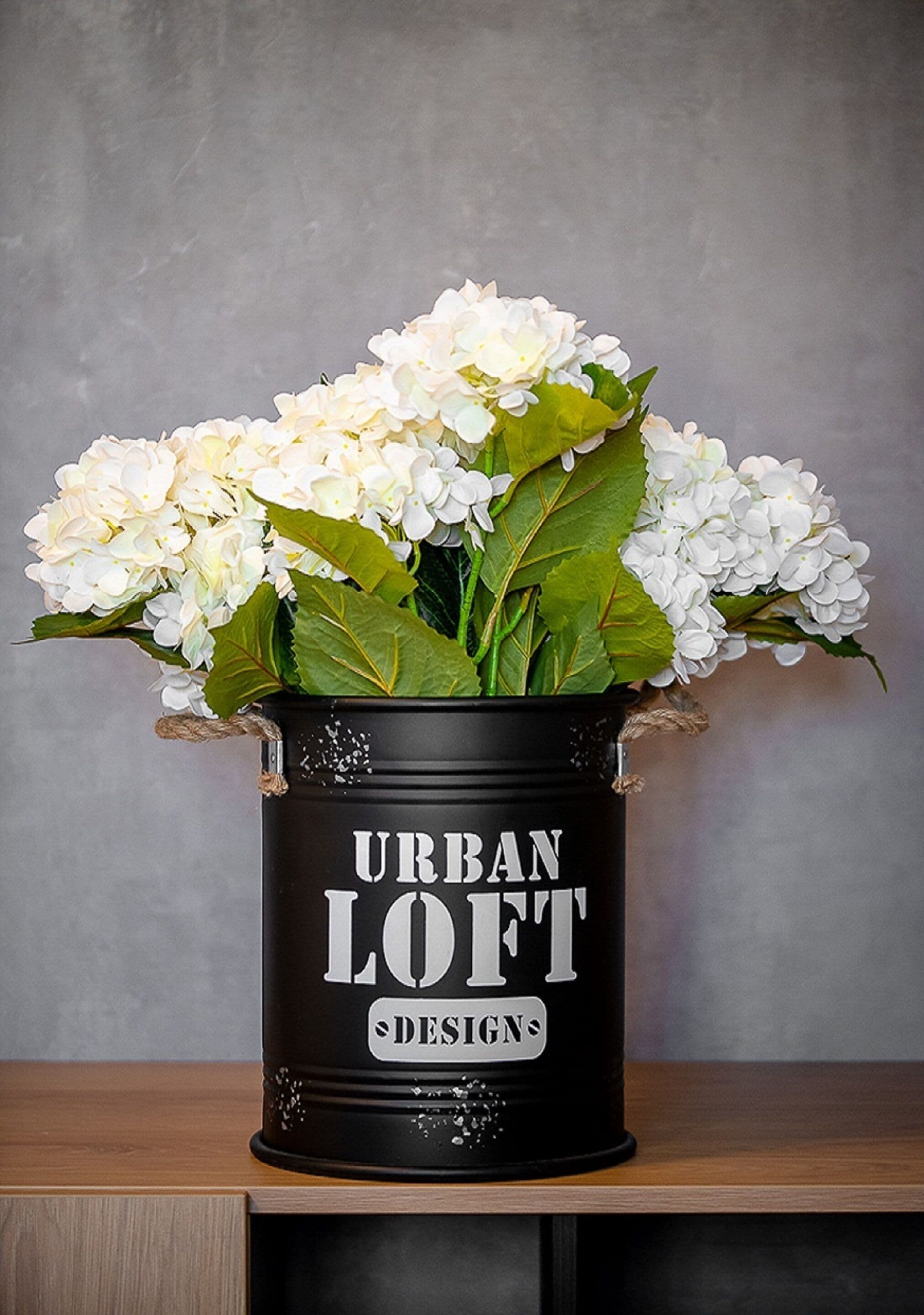 Een zwarte blik met de tekst 'URBAN LOFT DESIGNS', gevuld met witte bloemen en groene bladeren.