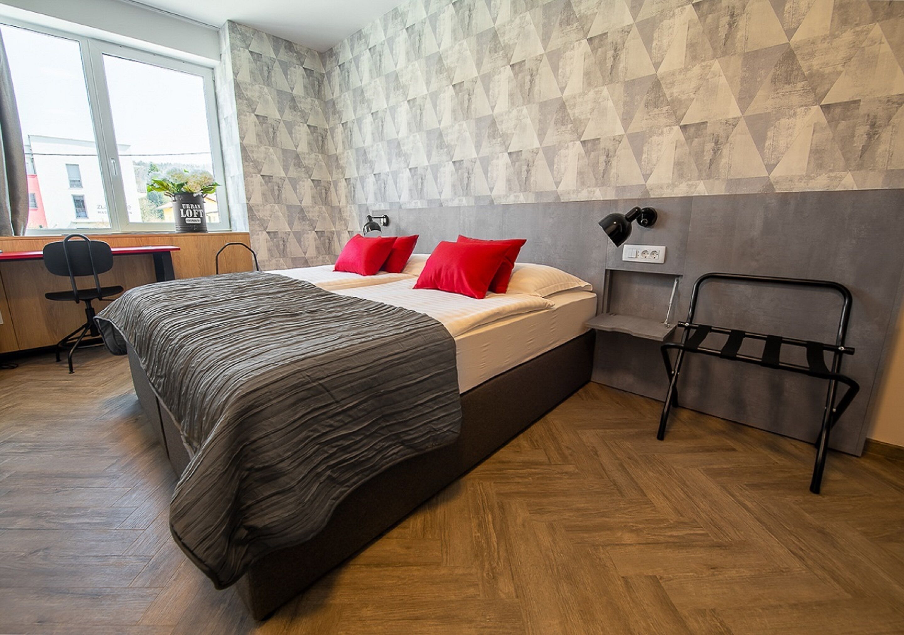 Ein modernes Schlafzimmer mit Bett, roten Kissen, einem Schreibtisch und einem Fenster.
