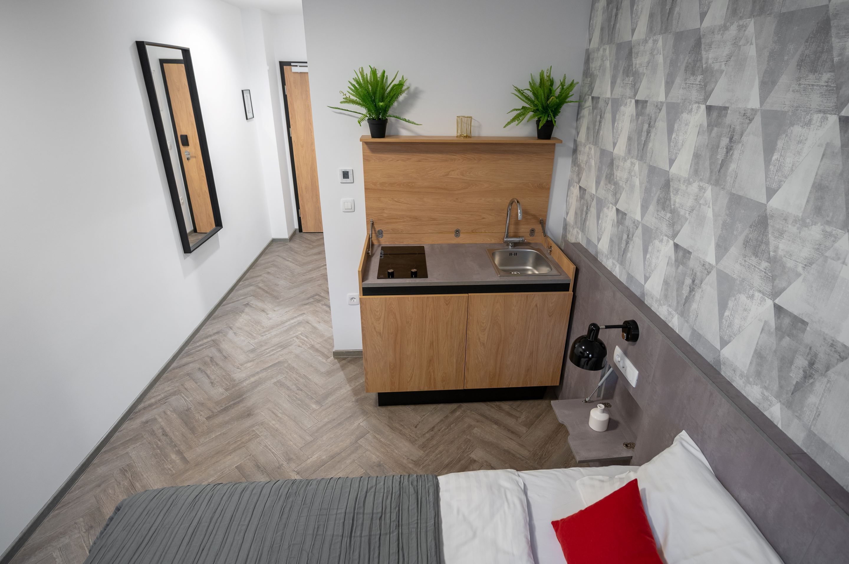 Ein kompaktes Hotelzimmer mit Küchenzeile, einem Bett und einem Parkettboden mit Fischgrätmuster.