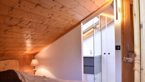 A bedroom with a sloped ceiling, a bed with white bedding, a bedside lamp, and a wardrobe with a glass door. - Meribel: Spacious 3BR Apartment, Sleeps 6, 150m from Ski Lift, Free Shuttle & Covered Parking (Méribel)