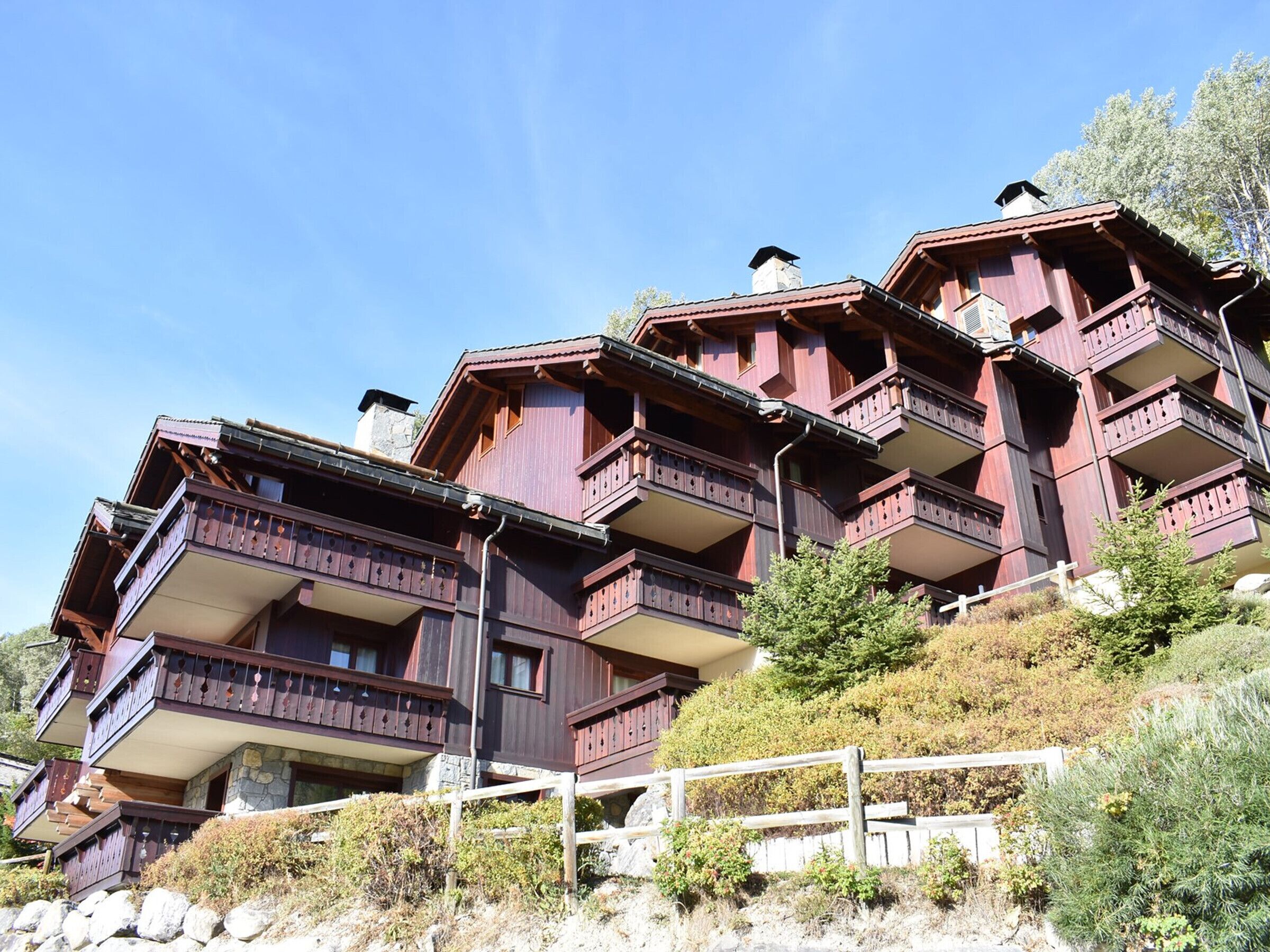 A multi-story wooden chalet with balconies, set on a hillside with a stone retaining wall and greenery.