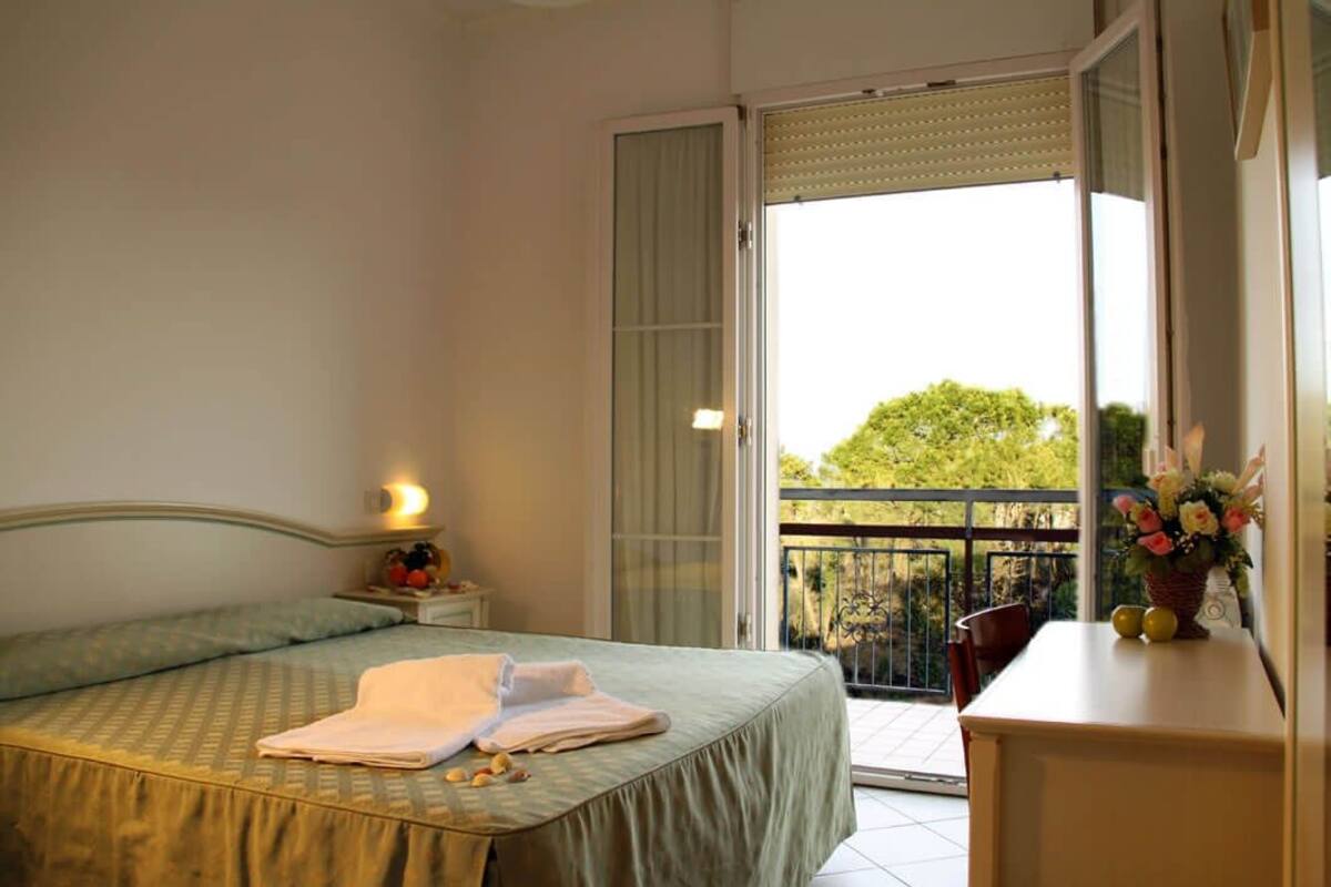 A bedroom with a bed, a nightstand, a window with a view of greenery, and a balcony with a railing.