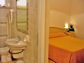 A bathroom with a sink, toilet, and mirror. There is a bed with an orange bedspread and a nightstand with a plant. - Ariston Vacanze Residence (Ravenna)