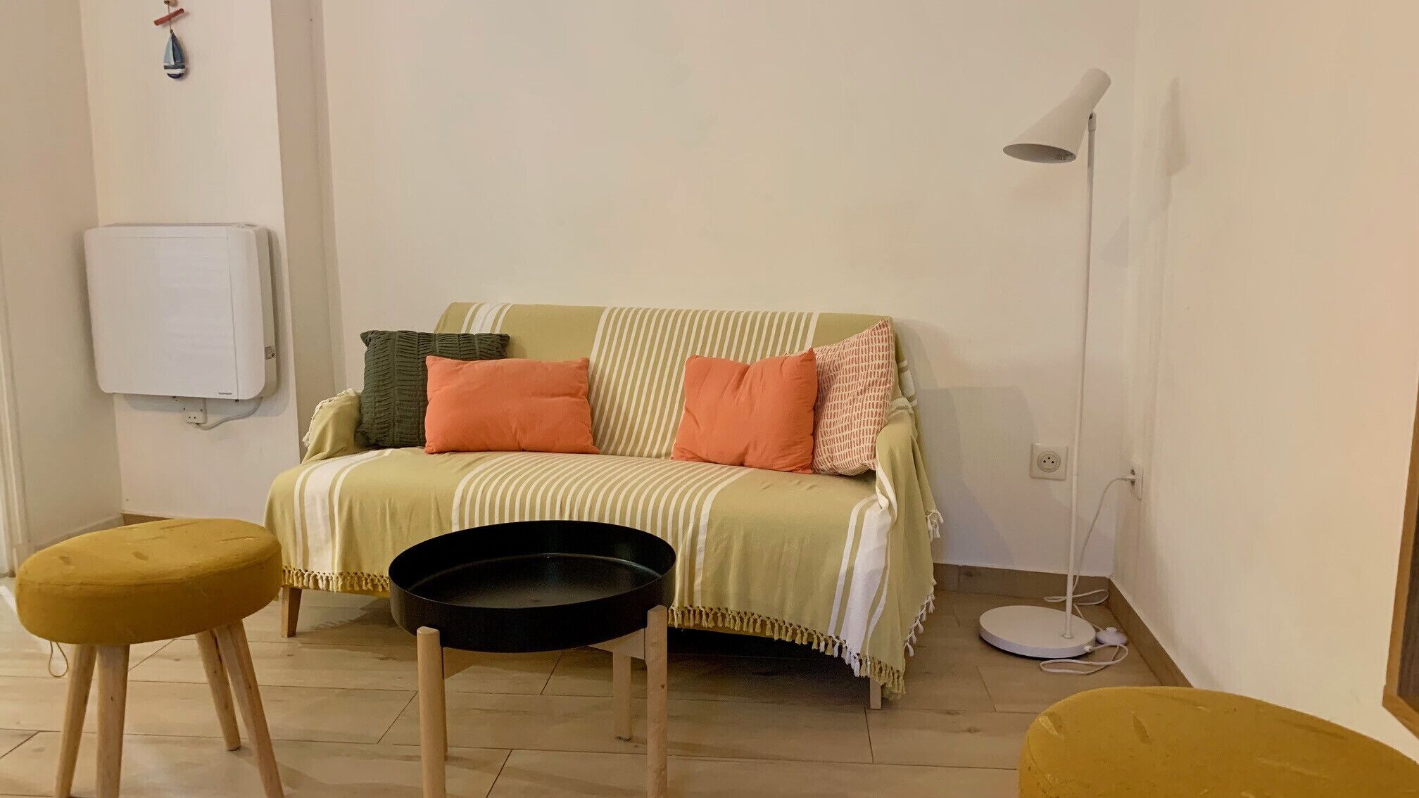 Una sala de estar acogedora con un sofá, dos taburetes, una lámpara de pie y un radiador.